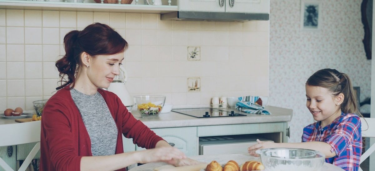 Young mother rolling dough and talking to her little cute daughter while cooking in the kitchen on weekend. Family, food, home and people concept