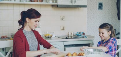 Young mother rolling dough and talking to her little cute daughter while cooking in the kitchen on weekend. Family, food, home and people concept